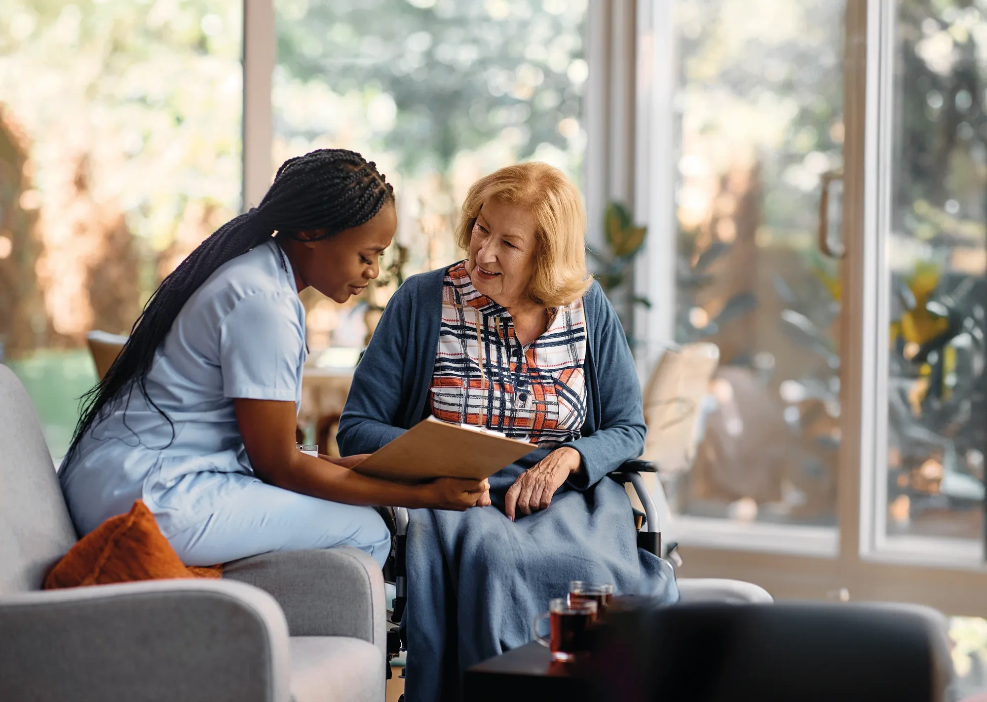 A care worker sitting beside an older woman in a wheelchair, going through paperwork with her in a sunlit room.