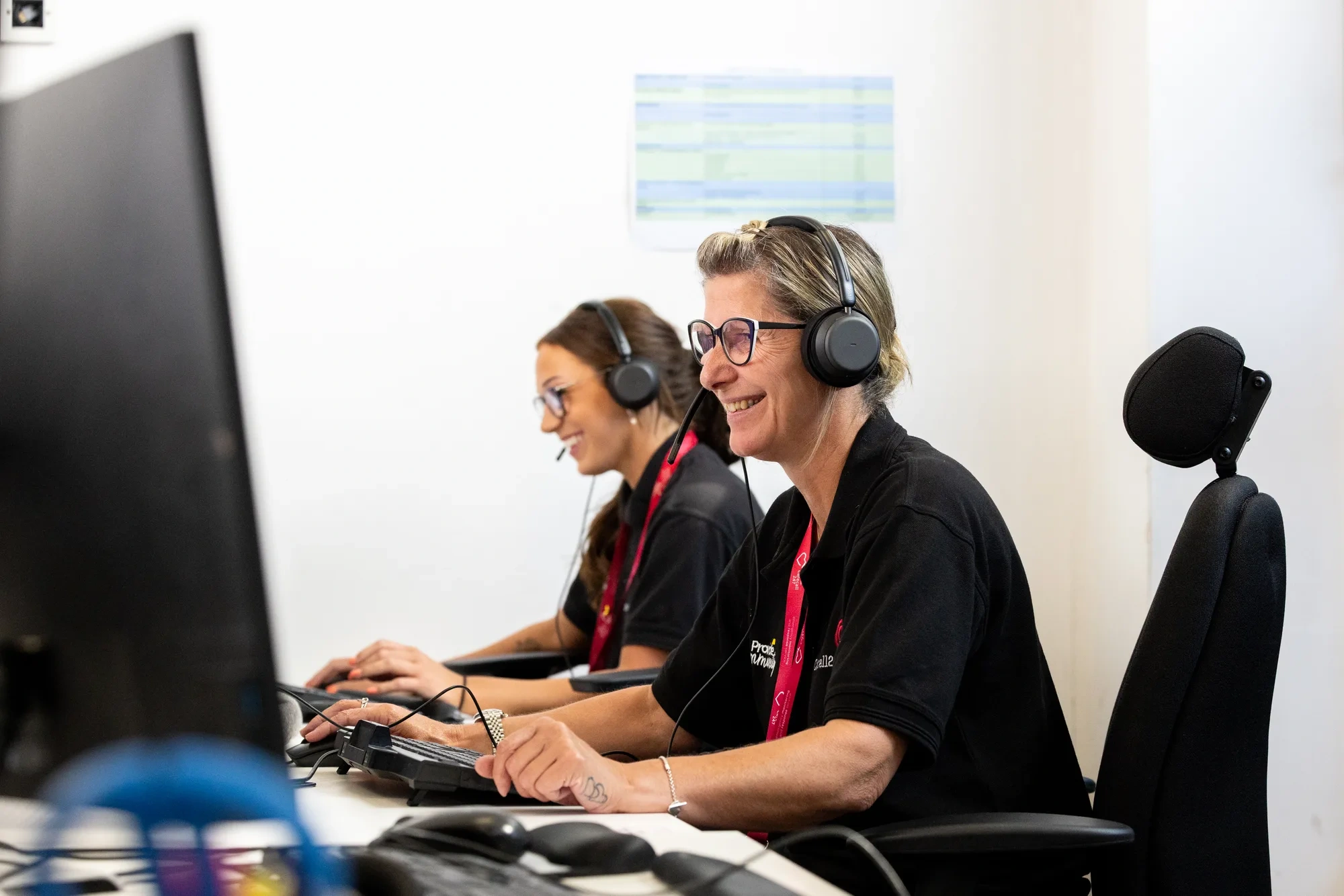 Two Provide Carecall247 monitoring centre operators wearing headsets at their workstations, one smiling as she takes a call.